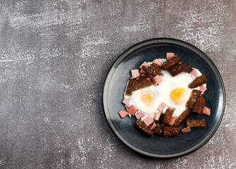 Fried eggs with dark rye bread and sausage on a round plate on a dark background. Top view, flat lay