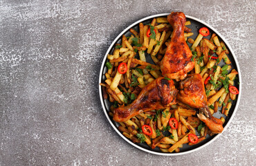 Chicken drumsticks with potatoes and hot peppers on a round plate on a dark background. Top view, flat lay