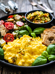 Continental breakfast - scrambled eggs, bread and greek salad on wooden table
