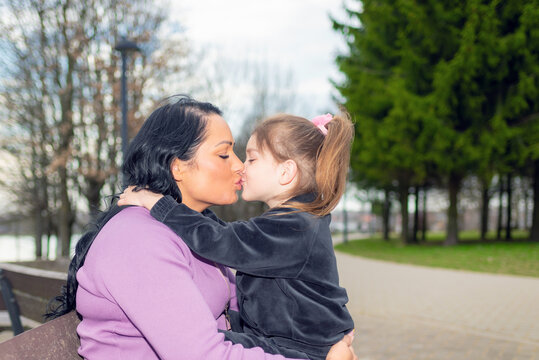 Cute 5 years old girl and her mom kissing at park on the bench in summer.Family and lifestyle concept.Closeup.