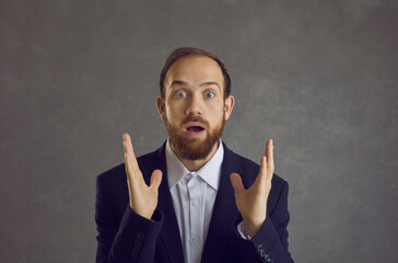 Adult shocked businessman entrepreneur in suit headshot portrait on grey background. Guy with widely opened eyes and mouth showing amazed funny facial emotion. People reaction and expression