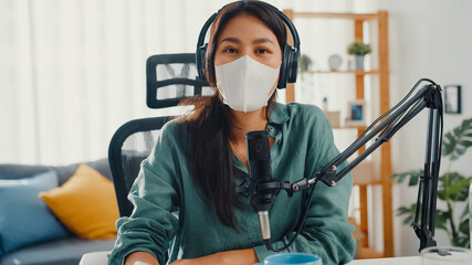 Teenager Asia girl record podcast use headphones and microphone wear mask protect virus look at camera talk in her room. Female podcaster make audio podcast from her home studio, Stay at home concept.