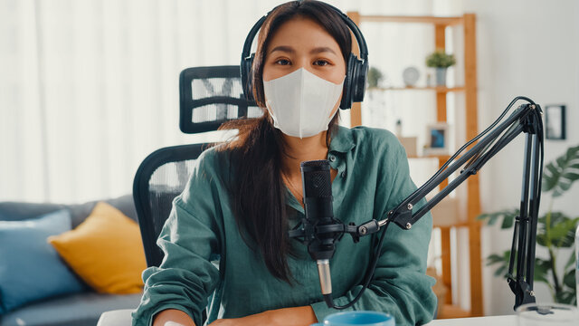 Teenager Asia girl record podcast use headphones and microphone wear mask protect virus look at camera talk in her room. Female podcaster make audio podcast from her home studio, Stay at home concept.