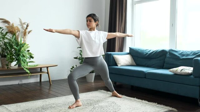 Young Indian woman does yoga at home bending knees to her chest in pindasana pose, standing on one leg doing an exercise for balance
