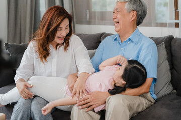 Obraz premium Asian grandparents talking with granddaughter at home. Senior Chinese, old generation, grandfather and grandmother using family time relax with young girl kid lying on sofa in living room concept.