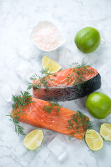 Raw salmon fillet slices with ice, fresh dill, limes and pink salt, vertical shot on a light-grey marble background