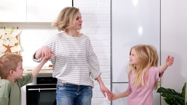 Excited Mom Or Babysiter And Two Kids Boy And Girl Jumping, Dancing Laughing In Modern Scandinavian House Kitchen. Happy Family Mother Daughter Son Having Fun Enjoy Playing Singing Together At Home.