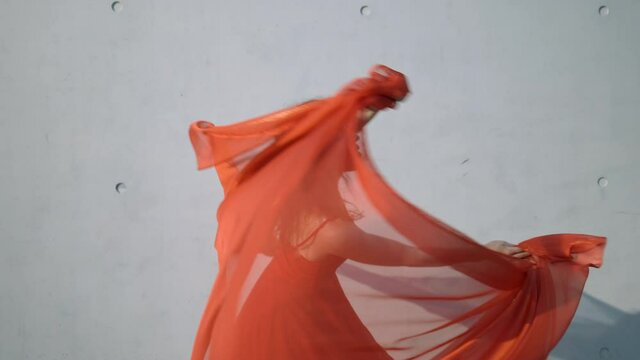 Slow Motion Of A Young Woman In A Orange Dress Dancing In The Sun With A Large Piece Of Orange Sheer Fabric And A Gray Concrete Wall In The Background - Berlin, Germany