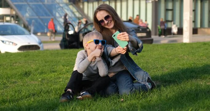 Mom and son laugh and take a selfie on a smartphone sitting on the lawn in the city. Mom and child have fun outdoors. Summer holidays