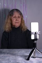 An elderly woman sits at a table in front of a smartphone on a tripod. Senior woman blogger is recording a video that is broadcast online indoors.