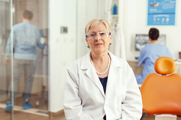 Obraz premium Portrait of specialist senior dentist woman in stomatological office while medical assistant discussing with patient in background. Stomatologist doctor looking on webcam