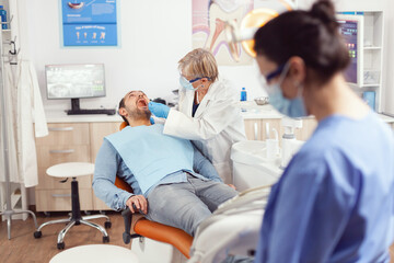 Obraz premium Senior stomatologist holding sterilized dental tools talking with sick patient before stomatological intervation. Nurse and doctor wearing masks cleaning teeth of man sitting on chair with open mouth