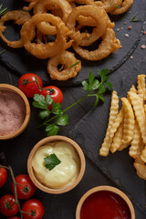 Homemade baked potato fries with mayonnaise, Tomato sauce and rosemary on stone board. fast food products : onion rings, french fries on cutting board, on black stone background, unhealthy food.
