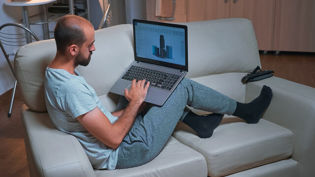 Tired arhitect man working at industrial cad prototype while sitting on sofa. Caucasian male looking at laptop computer display for new technical construction concept late at night in kitchen