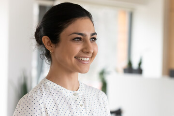 Three quarter portrait of smiling confident attractive Indian businesswoman standing in office, successful young woman entrepreneur employee executive looking to aside, business vision concept