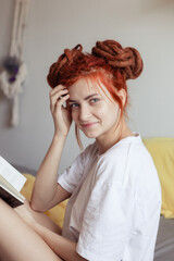 Very pretty redhead girl with dreads on bed in bedroom in the morning. Young woman reading a book sitting in bed. A feminine, gentle girl.