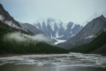 Atmospheric alpine landscape with mountain lake with streams from snowy mountains in overcast weather. Gloomy mountain scenery with green lake with rainy circles and low clouds in mountain valley.