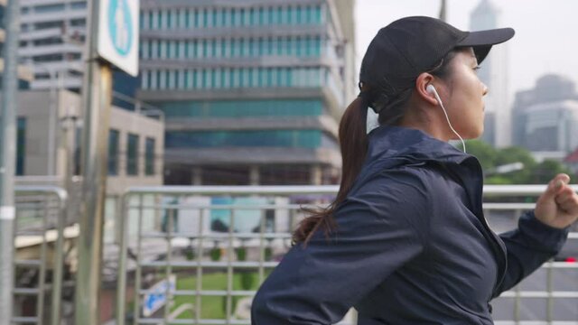 Asian woman in sportswear jogging in the city in summer morning. Athlete female enjoy healthy outdoor lifestyle do sport training workout exercise fitness running with listen to music from earphones.