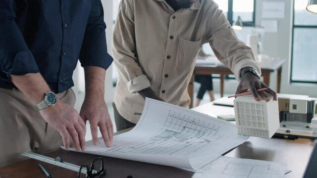 Tilt up shot of senior Caucasian architect unrolling floor paper plan on office table and discussing it with young Afro-American male coworker