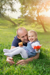 Naklejka na ściany i meble A little girl is eating a pie in her father's arms and laughing merrily. The relationship between father and daughter. Picnic in the park at sunset. A loving father. Happy family. Childhood in harmony