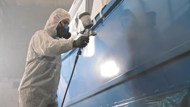 Low angle medium long of worker wearing white protective suit with hood and respirator, spraying blue paint on boat in progress