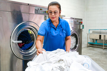 A laundry staff wearing a blue uniform is checking towels from a laundry trolley in front of industrial washing machines. Shot taken in the factory. - obrazy, fototapety, plakaty