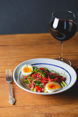 Tuna salad with vegetables, boiled eggs, cherry tomatoes and spices and one glass of red wine over wooden background.