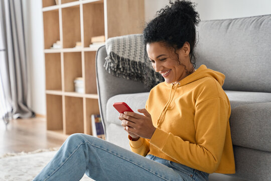 Happy smiling young African American mixed raced girl holding smartphone sitting on floor at home using online entertainment mobile applications surfing social media searching internet.