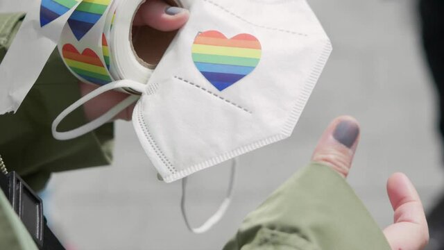 Close up of hands with painted nails placing a lgbtq rainbo heart shaped sticker on an ffp2 mask during a pride demonstration on a sunny day. Symbol of lgbtq+, gay, lesbian, transgender rights 4K