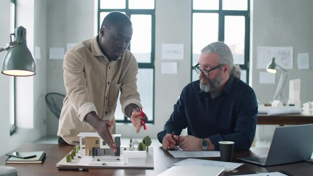 Young black male architect discussing building model with senior colleague while working together on project in office