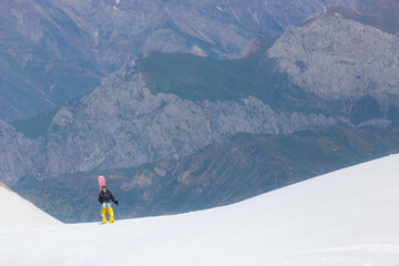 Snowboarder walks through the snow against the backdrop of green mountains