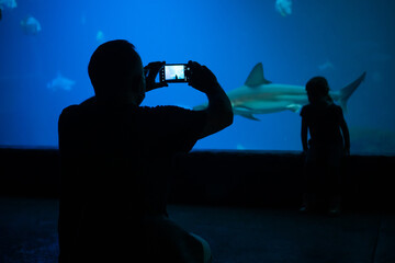 Father make a photo of daughter standing in front of aquarium with shark and other fishes inside.