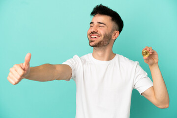 Fototapeta premium Young man holding a Bitcoin isolated on blue background giving a thumbs up gesture