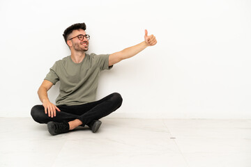 Young caucasian man sitting on the floor isolated on white background giving a thumbs up gesture