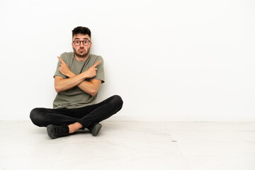 Young caucasian man sitting on the floor isolated on white background pointing to the laterals having doubts