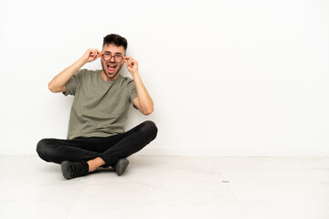 Fototapeta premium Young caucasian man sitting on the floor isolated on white background with glasses and surprised