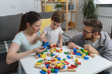 Mother and father playing with their cute smart toddler son at home using colorful wooden toys