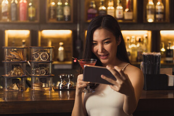 Asian woman taking a photo of herself while drinking whiskey at the bar.