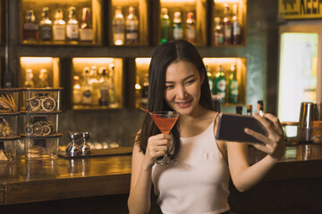 Asian woman taking a photo of herself while drinking whiskey at the bar.