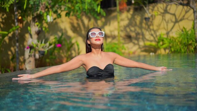 Smiling happy Asian lady in a swimming pool wearing a black bikini and white sunglasses in Tropical hotel. She is raising and stretching her arms up, slow-motion