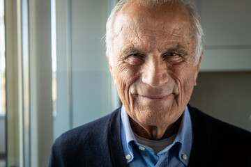 Cinematic shot of happy senior pensioner man is smiling in camera in living room at home. Concept of life, family, achievement, father, husband, grandfather, elderly, retirement, boarding house.