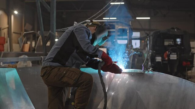 Medium long of unrecognizable dockyard worker wearing helmet, welding new metal boat indoors