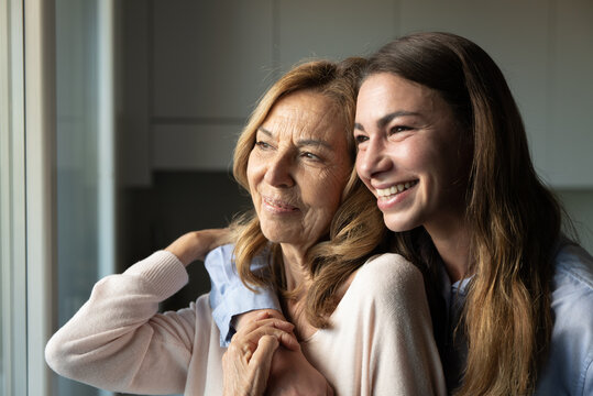Cinematic shot of happy granddaughter is hugging her grandmother as sign of love and respect while looking through window at home. Concept: life, grandparents, generation, love, care, family.
