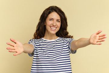 Cheerful curly female with happy smile hold open hands raised for embrace. Give me a hug. Portrait of joyful glad young woman with outstretched arms for hugging isolated over beige studio background