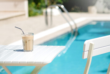 Ice coffee Cyprus Frappe Fredo against blue clear water of the swimming pool, on white table, with metal straw . Summer minimalistic background, holiday or vacation concept. Sun and shadows.Copy space