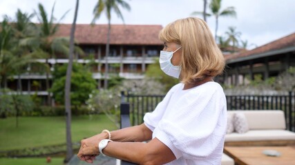 Fototapeta na wymiar Side view of a middle aged woman wearing a face enjoying her view from a balcony. Tropical holidays during covid-19 pandemic