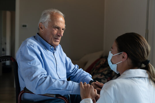 Cinematic Shot Of Social Worker With Medical Mask Stroking Hands Of Senior Man As Sign Of Care And Respect In Wheelchair At Home. Concept Of Healthcare, Protection, Assistance, Caregiver, Covid-19.