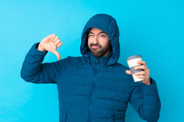 Man wearing winter jacket and holding a takeaway coffee over isolated blue background showing thumb...
