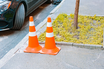 Two orange traffic cones with white stripes stands on a sidewalk near tree and ground. Security....
