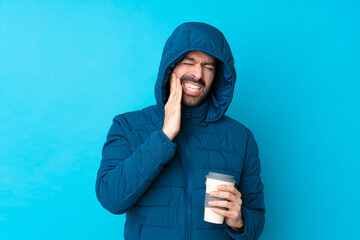 Man wearing winter jacket and holding a takeaway coffee over isolated blue background with toothache